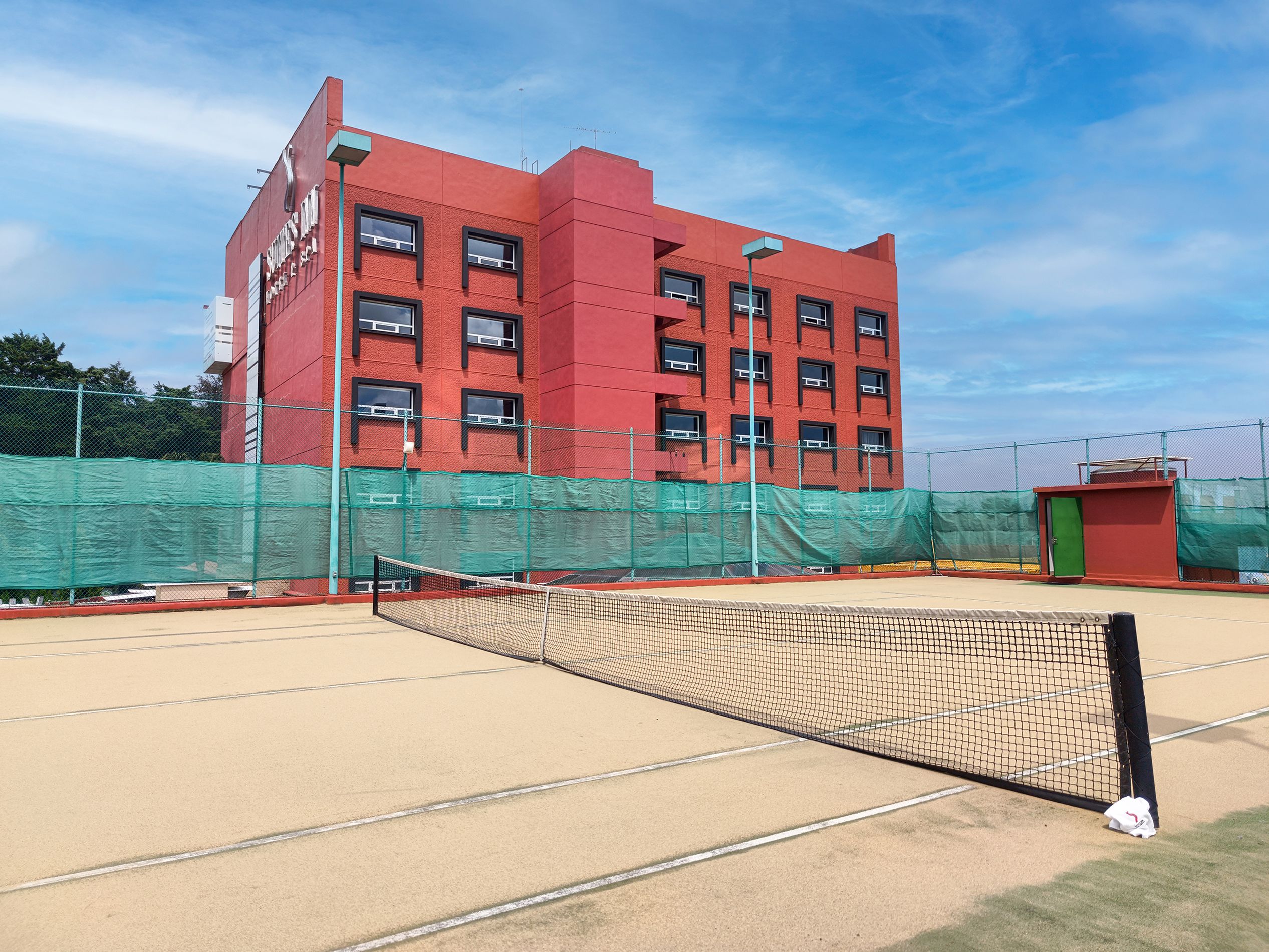 A tennis court with a net, surrounded by a green fence, and a red building in the background.