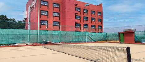 A tennis court with a net, surrounded by a green fence, and a red building in the background.