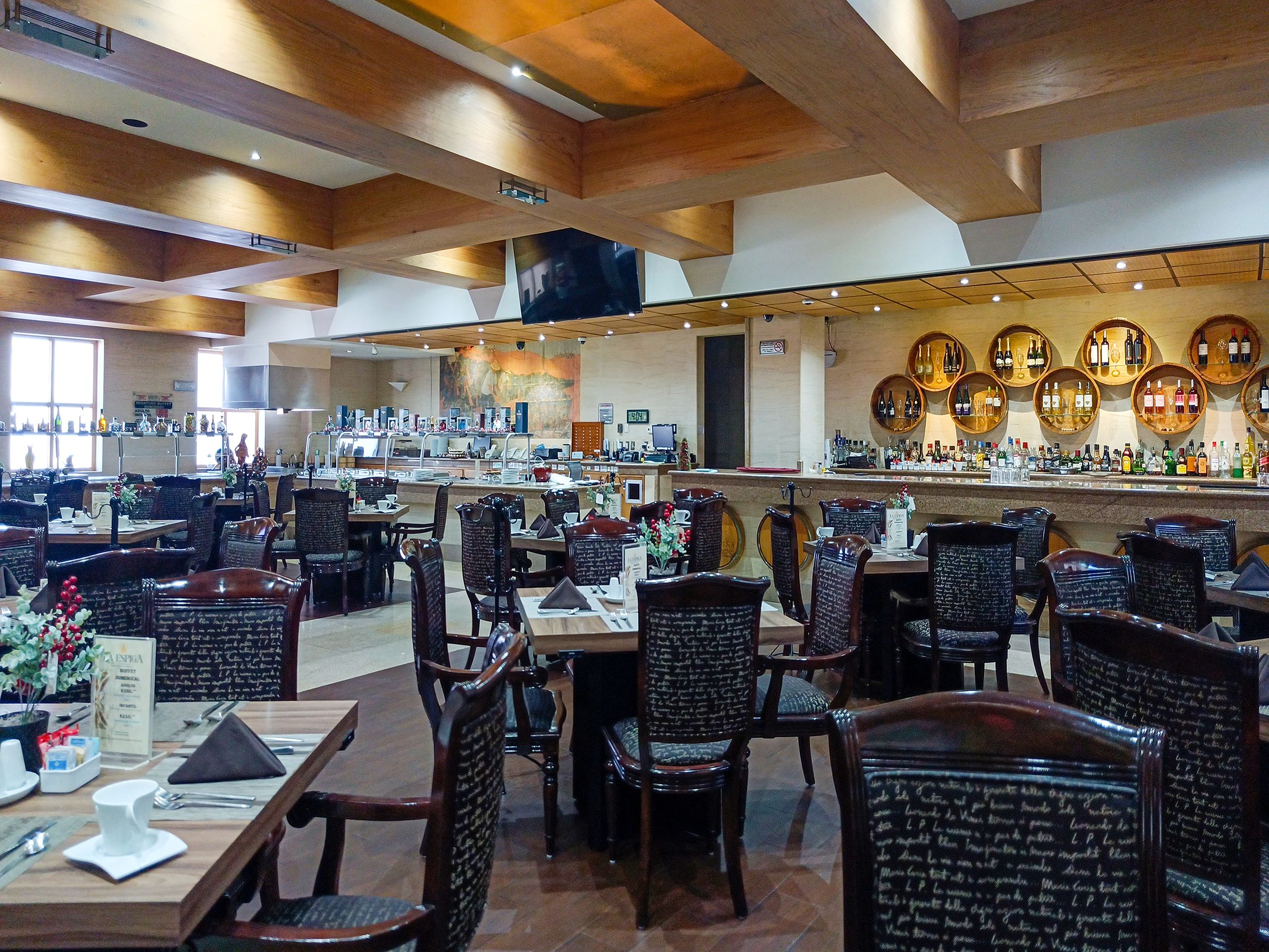 A restaurant interior with multiple dining tables and chairs, a bar area with bottles, and a large mural on the wall.