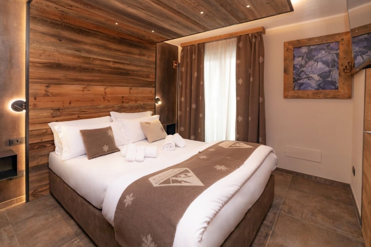 A modern bedroom with a wooden headboard, a large bed with white and brown bedding, a painting on the wall, and a small shelf with a lamp.