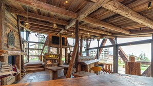 Un interior rústico con vigas de madera a la vista, ventanas amplias con vistas a edificios y follaje, y una zona de bar con taburetes.