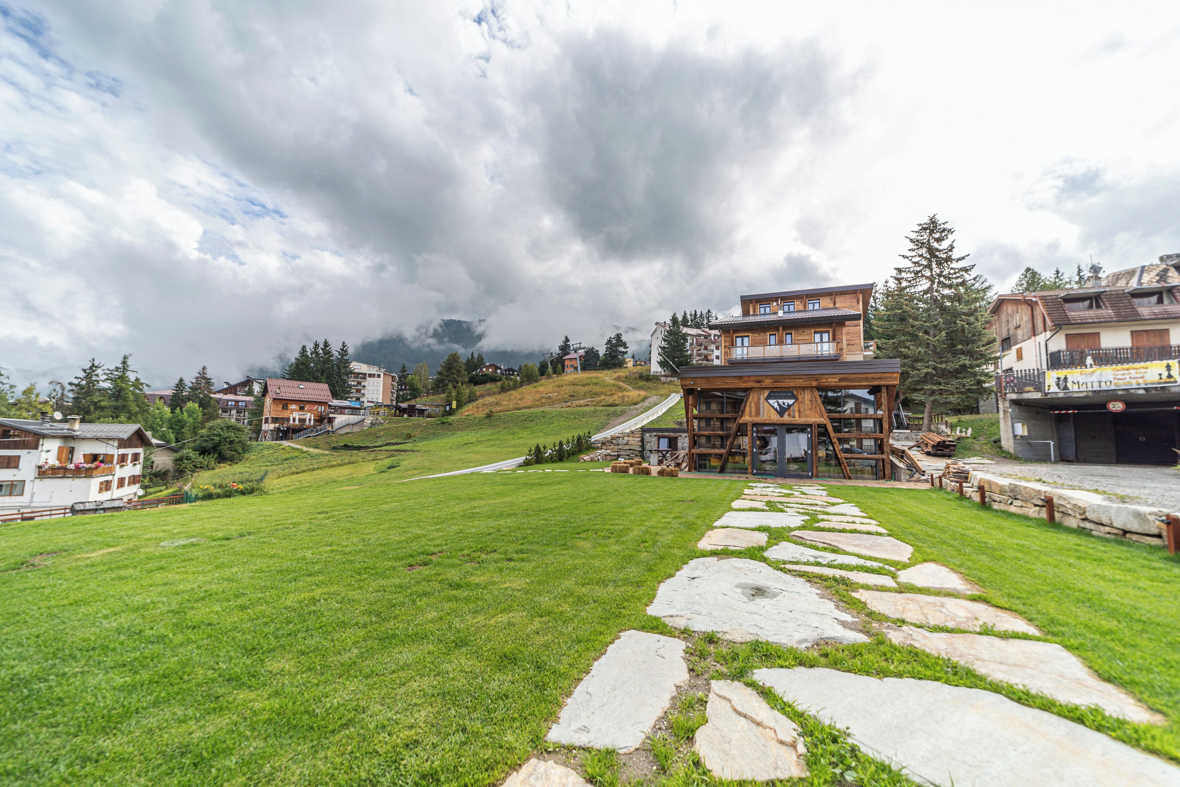Un chalet rustique avec un sentier de pierres menant à une construction en bois, entouré de verdure et de maisons sur une colline.