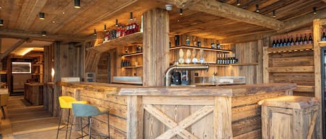 Un rústico espacio de bar con vigas de madera, mostrador, taburetes y estantes llenos de botellas.