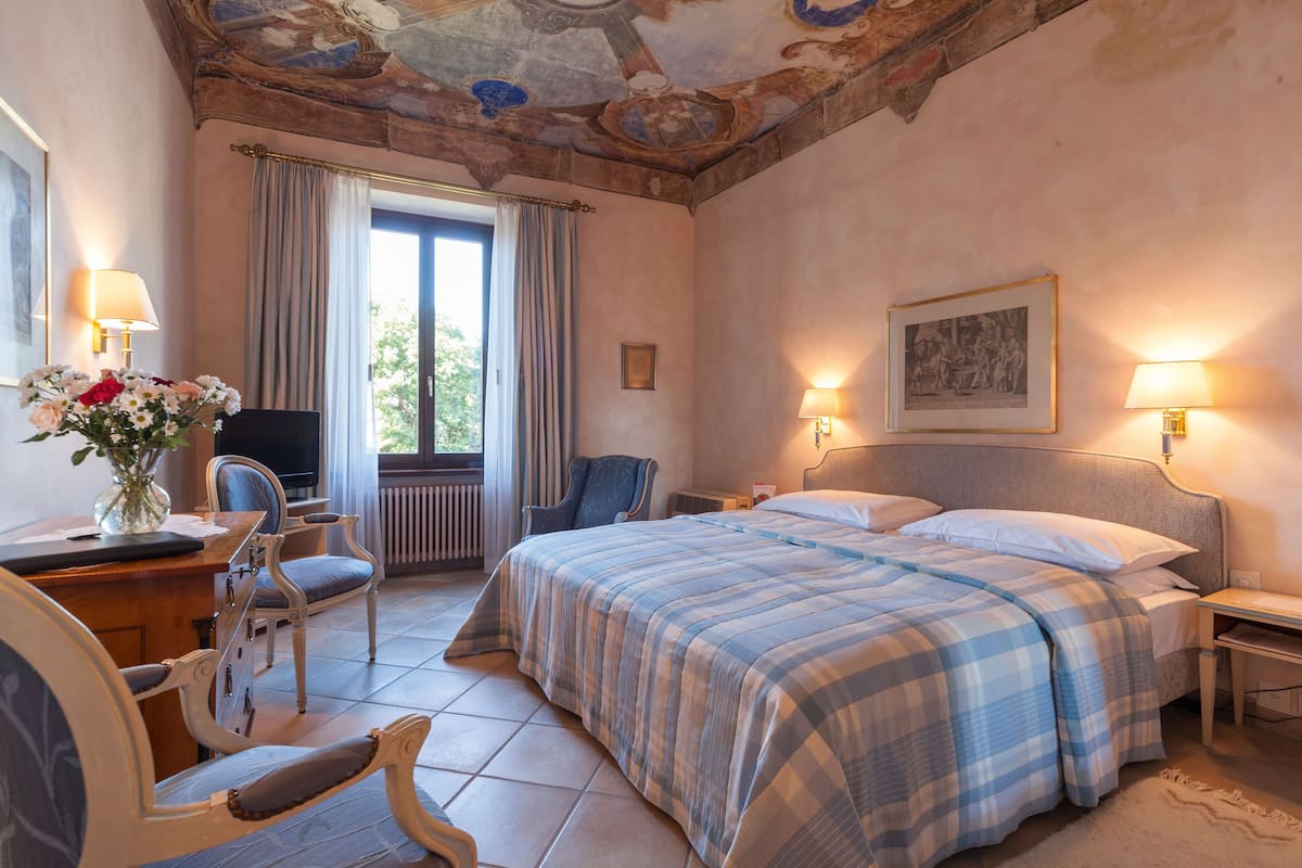 A bedroom with a bed, two bedside tables, a desk with a chair, a TV, and a window with curtains.