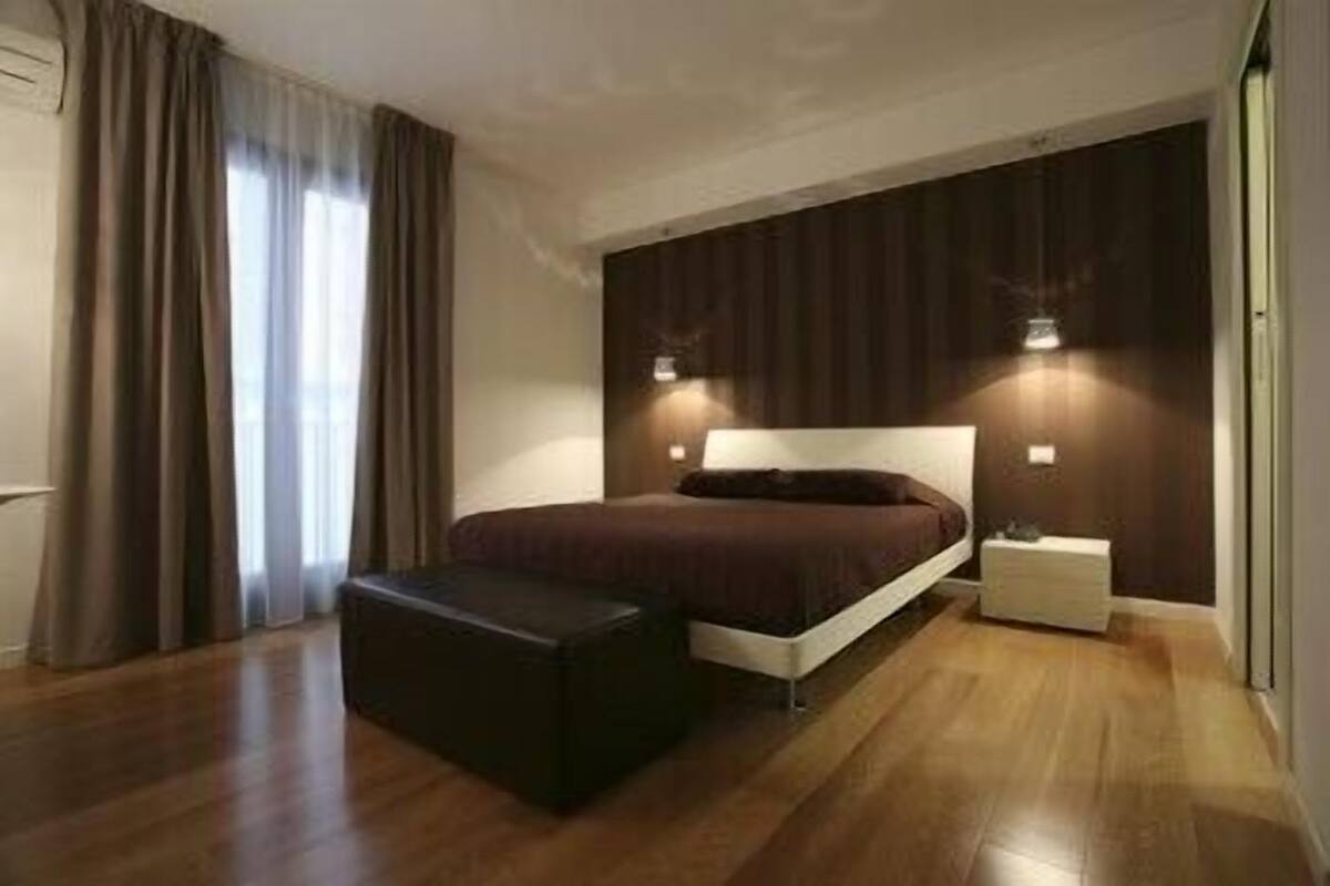 A modern bedroom with a wooden floor, a large bed with a dark headboard, a bedside table, and a small nightstand.