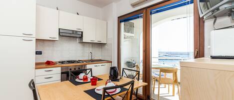 A compact kitchen with white cabinets, a wooden table, and chairs. A large window offers a view of the sea.