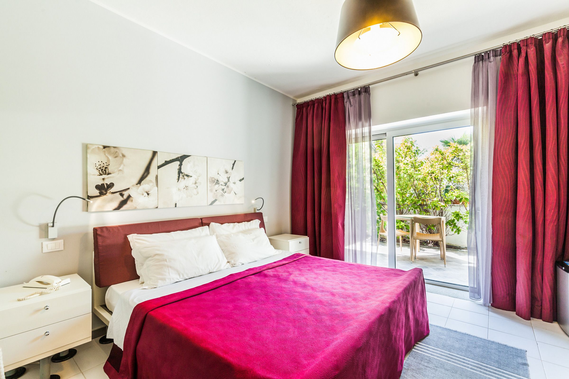 A hotel room with a large bed, a nightstand with a phone, a wall-mounted lamp, and a view of the outdoors through a sliding glass door.