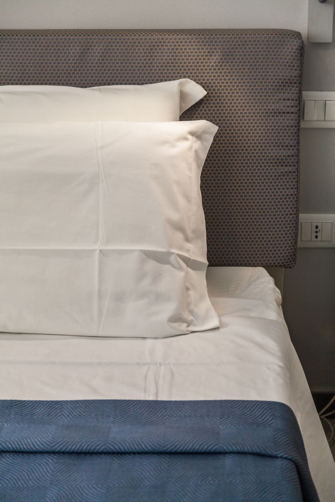 Ein ordentlich bezogenes Bett mit weißer und blauer Bettwäsche, ein grauer Kopfteil mit strukturierter Oberfläche und eine Steckdose an der Wand.