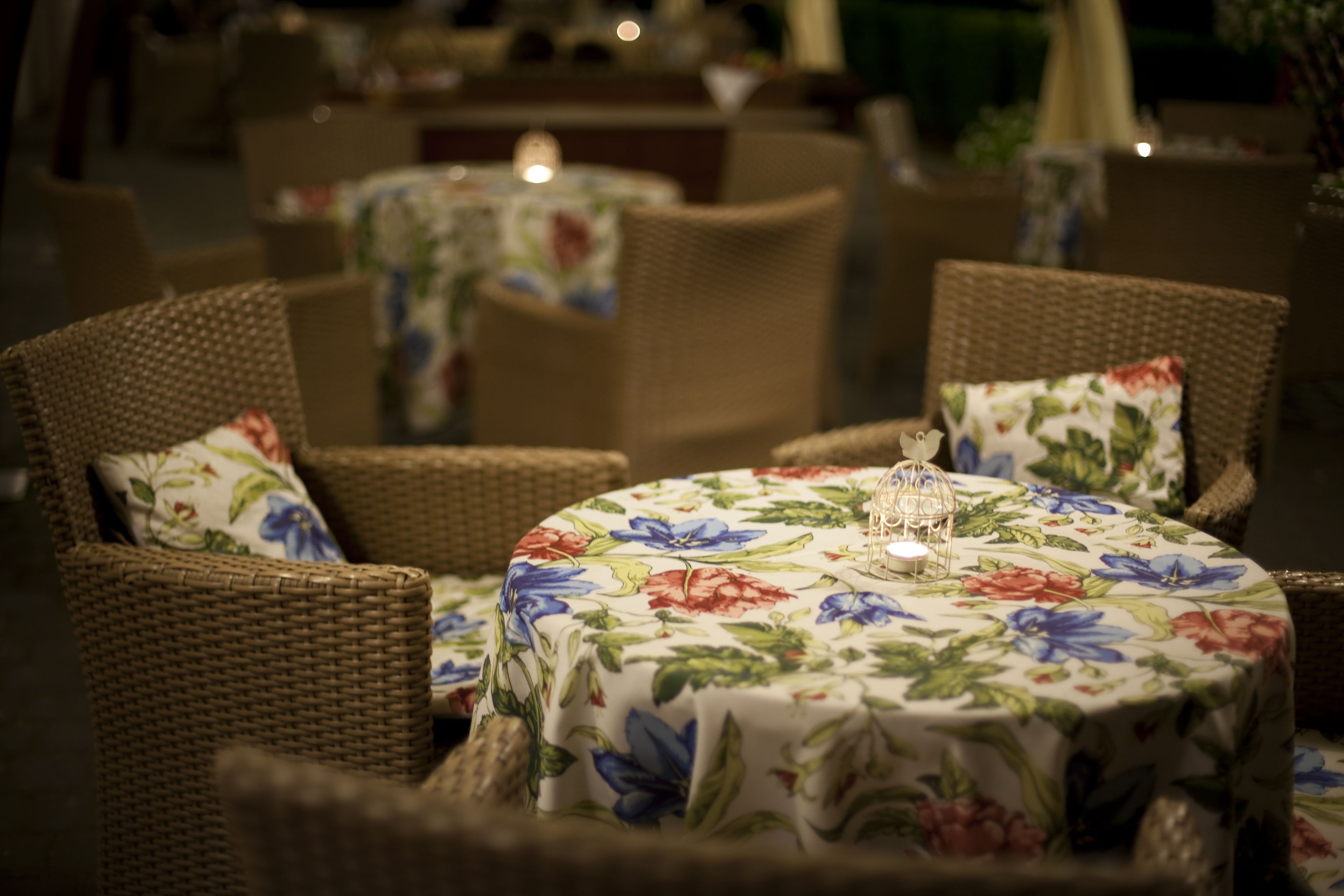 A dining area with wicker chairs and round tables covered with floral tablecloths.