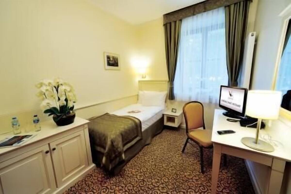 Une chambre d’hôtel comprenant un lit, un bureau avec un ordinateur, une chaise, une petite table avec une lampe, une table de chevet, une table de nuit, une plante en pot et une fenêtre avec des rideaux.