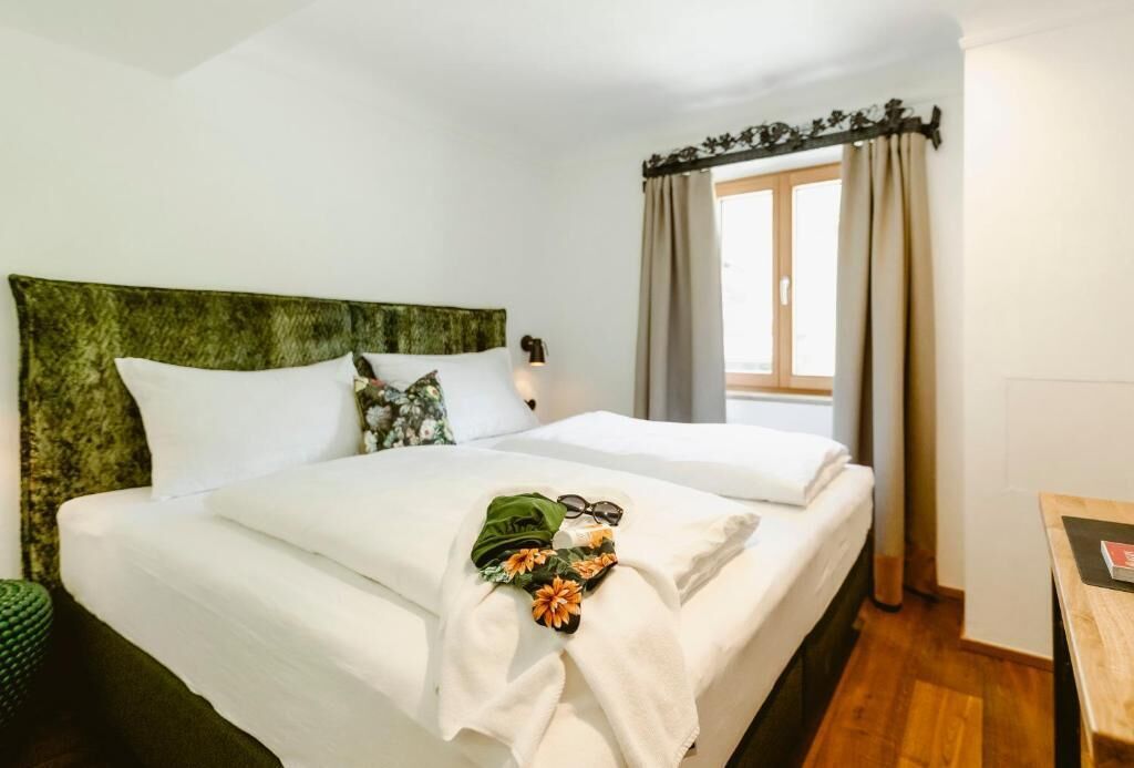 A hotel room with a bed, a window with curtains, a bedside table, and a green headboard.