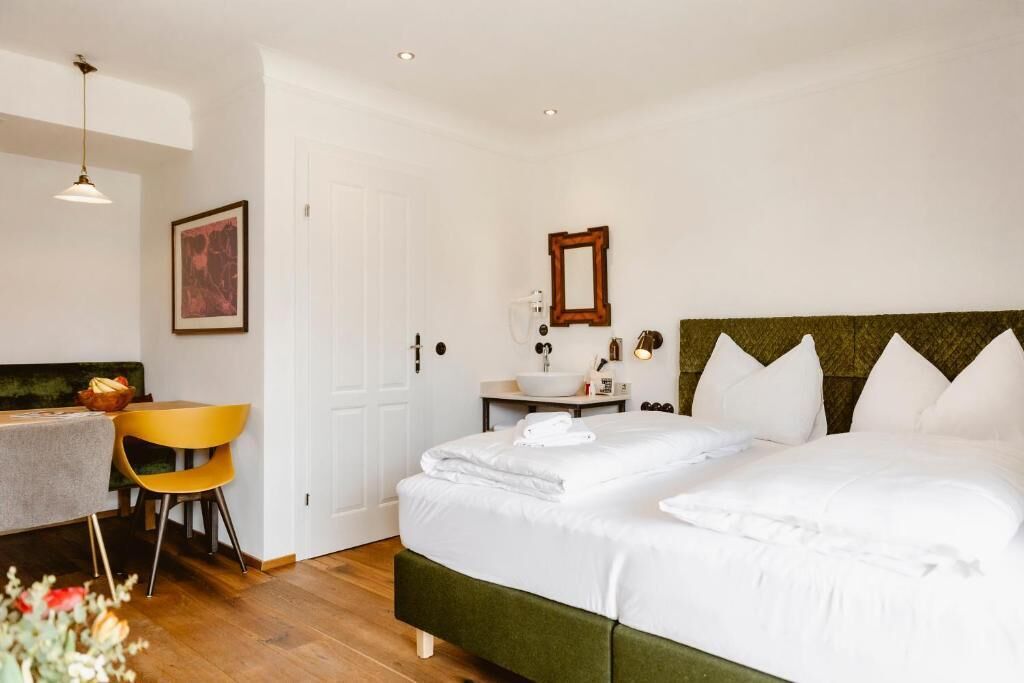 A hotel room with a bed, a bedside table, a chair, a desk, a sink, and a mirror.