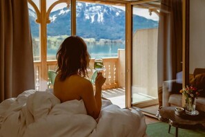 A person sitting on a bed with a view of a mountain and lake through a glass door. - Berau Landhotel am See (Sankt Wolfgang im Salzkammergut)
