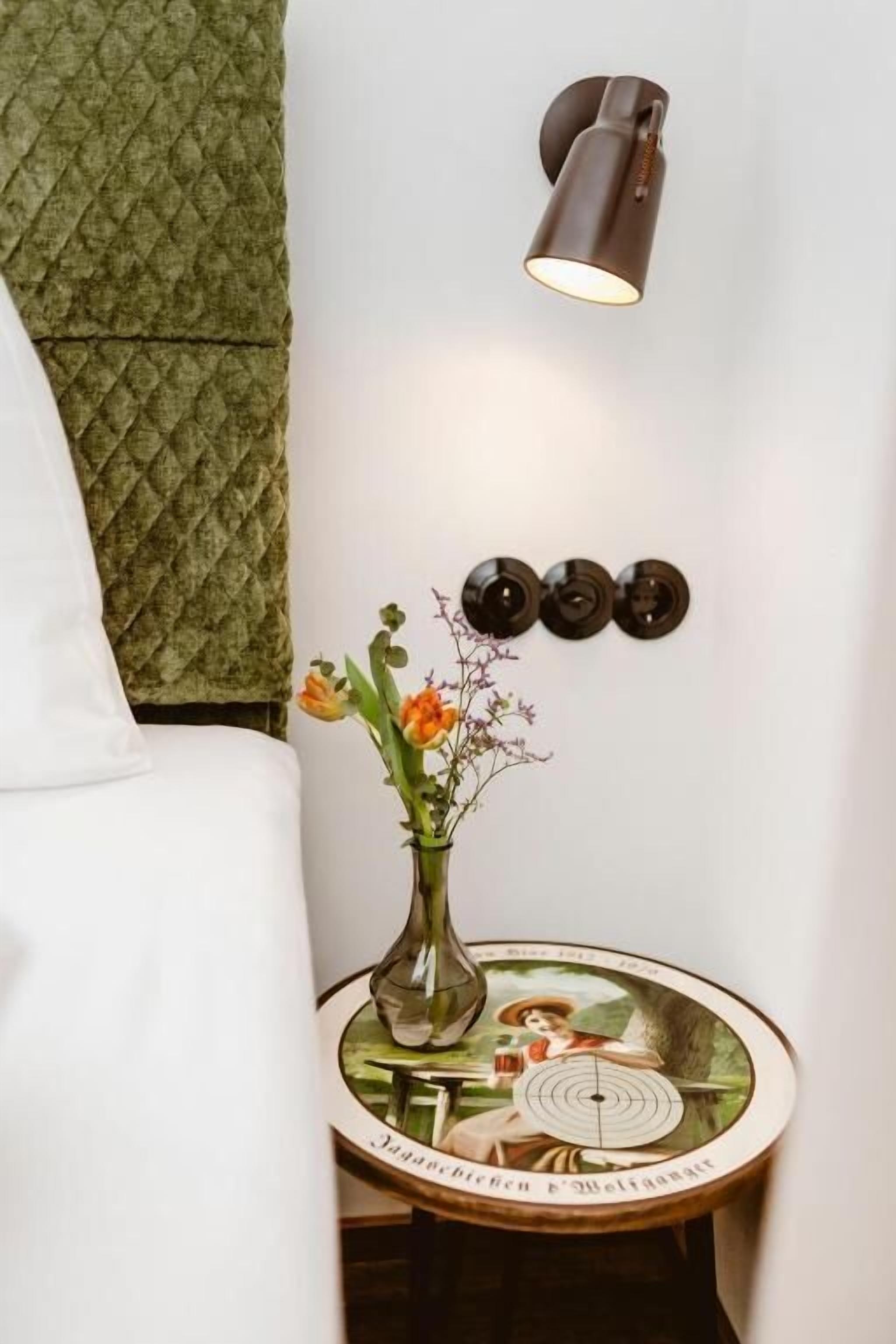 A bedside table with a vase of flowers, a wall-mounted lamp, and a decorative plate.
