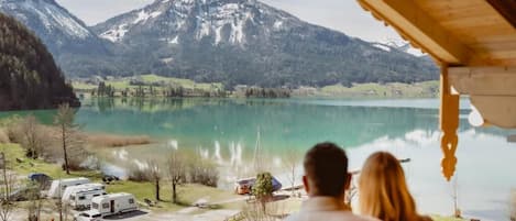 Una pareja contempla un lago con montañas al fondo, un área techada con vistas y vehículos recreativos estacionados cerca.