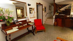 A reception area with a wooden desk, a red chair, a vase with flowers, a mirror, and a staircase leading to a landing with bookshelves. - Albergo Ristorante Umbria (Città di Castello)