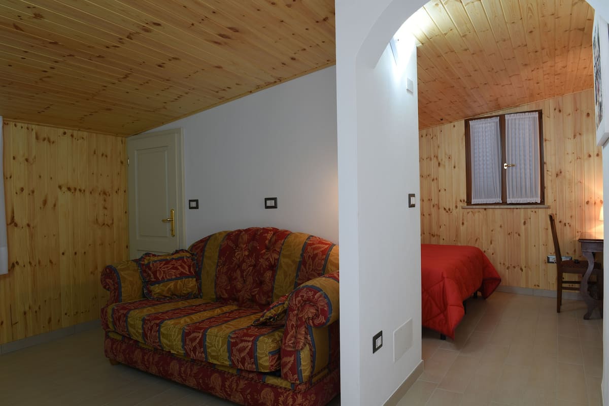 A room with wooden walls and ceiling, a red sofa, a bed with a red cover, and a wooden chair.