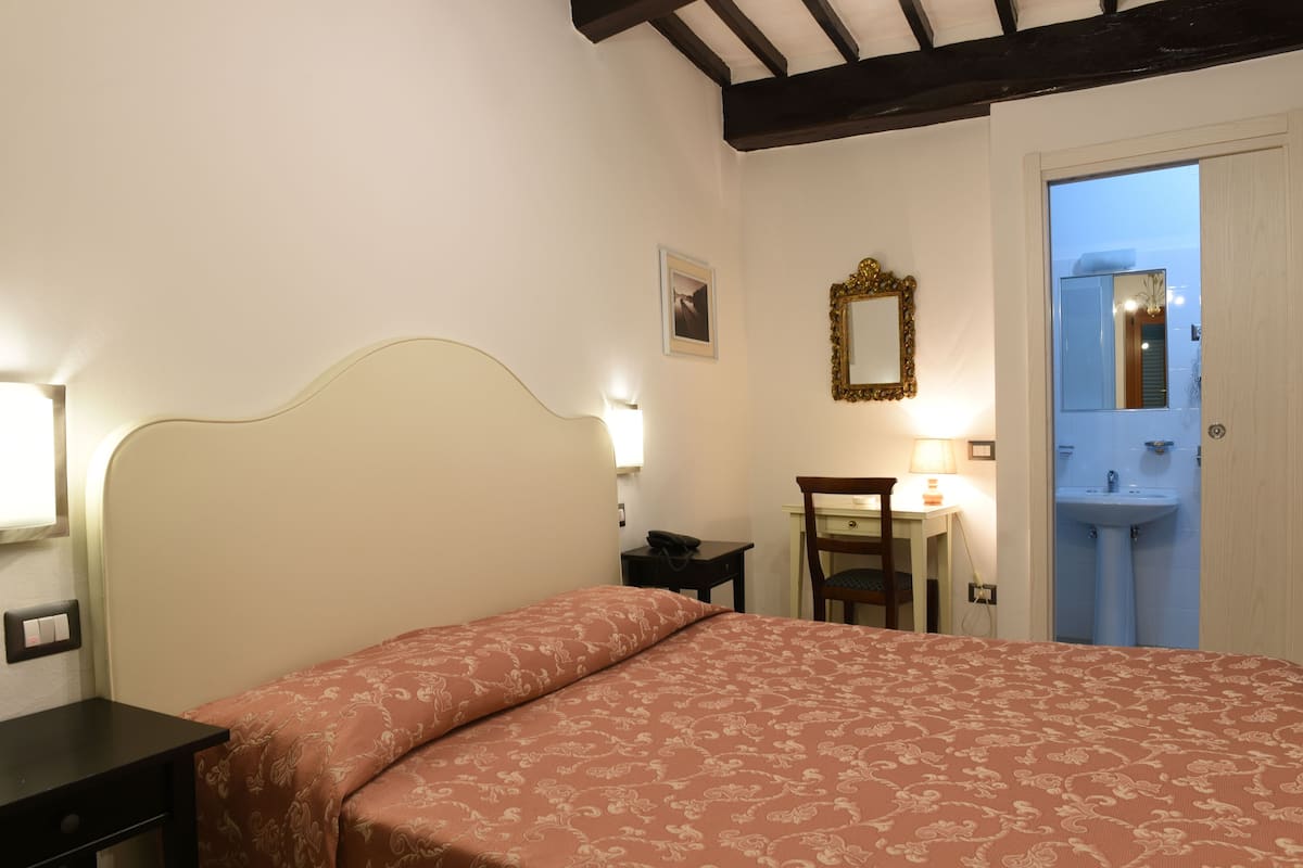 A bedroom with a bed, bedside table, lamp, and a bathroom visible through an open door.