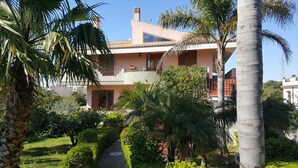 A two-story house with a balcony, surrounded by lush greenery and palm trees. - Villa Ottavia (Noto)