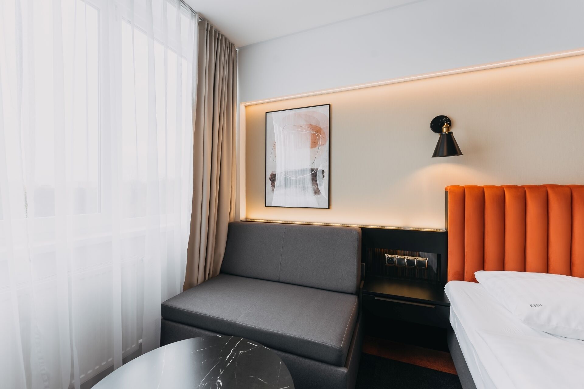 A hotel room with a grey sofa, a small round table, a bed with white bedding, an orange headboard, a black lamp, and a framed abstract artwork on the wall.