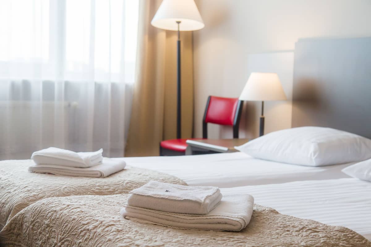 Une chambre d’hôtel avec un lit bien fait, deux lampes de chevet, un fauteuil rouge et une fenêtre ornée de rideaux légers.