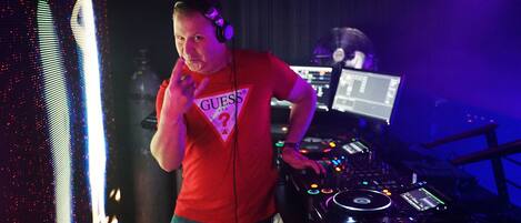 A DJ with headphones and a Pioneer DJ controller in a dimly lit room with a large screen displaying vertical lines.