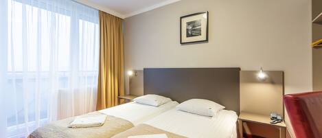 Quarto de hotel com duas camas, uma escrivaninha, uma cadeira, mesa de cabeceira, luminária e um quadro na parede.