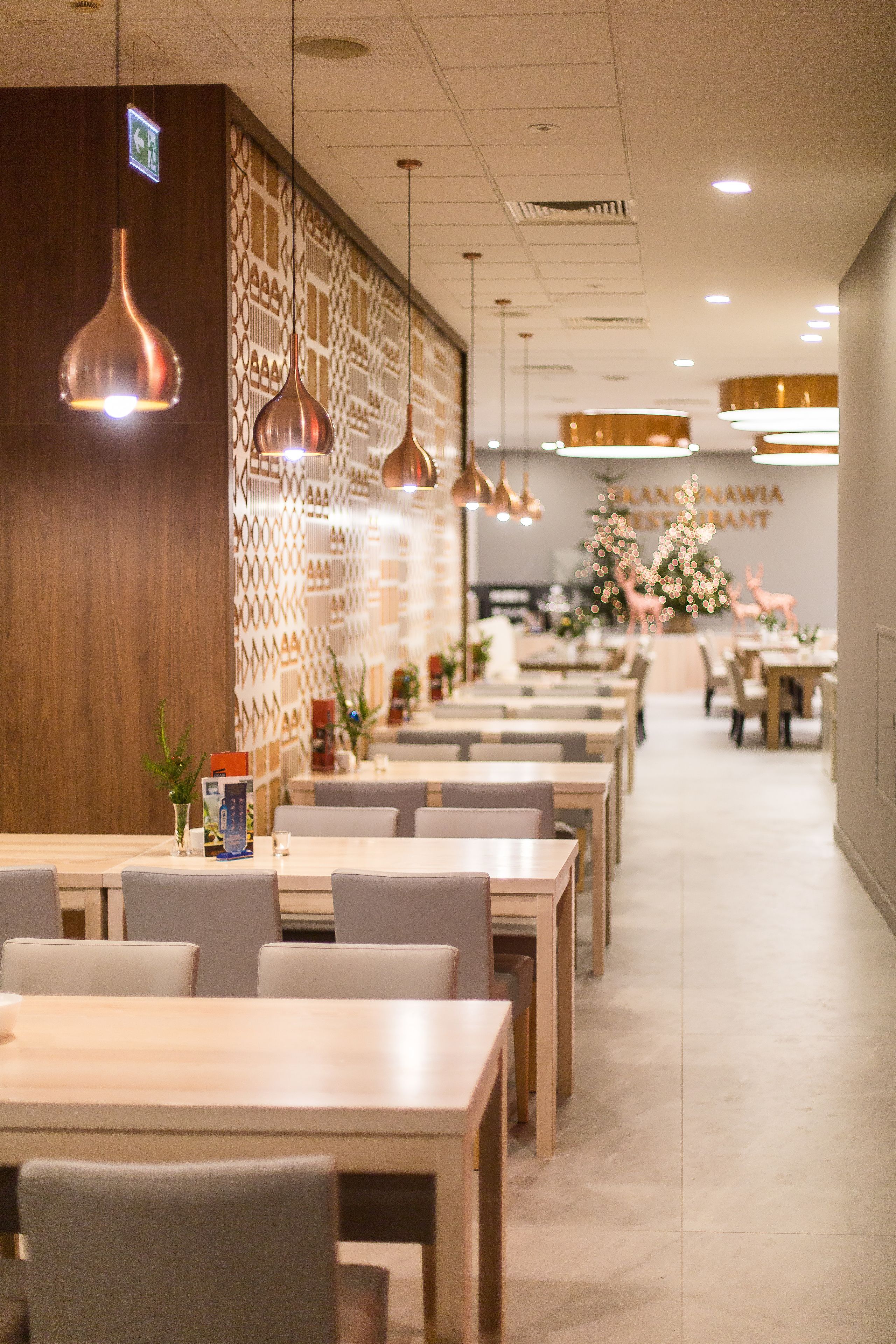 Un intérieur de restaurant moderne avec de longues tables, des suspensions en cuivre et un mur orné de motifs.