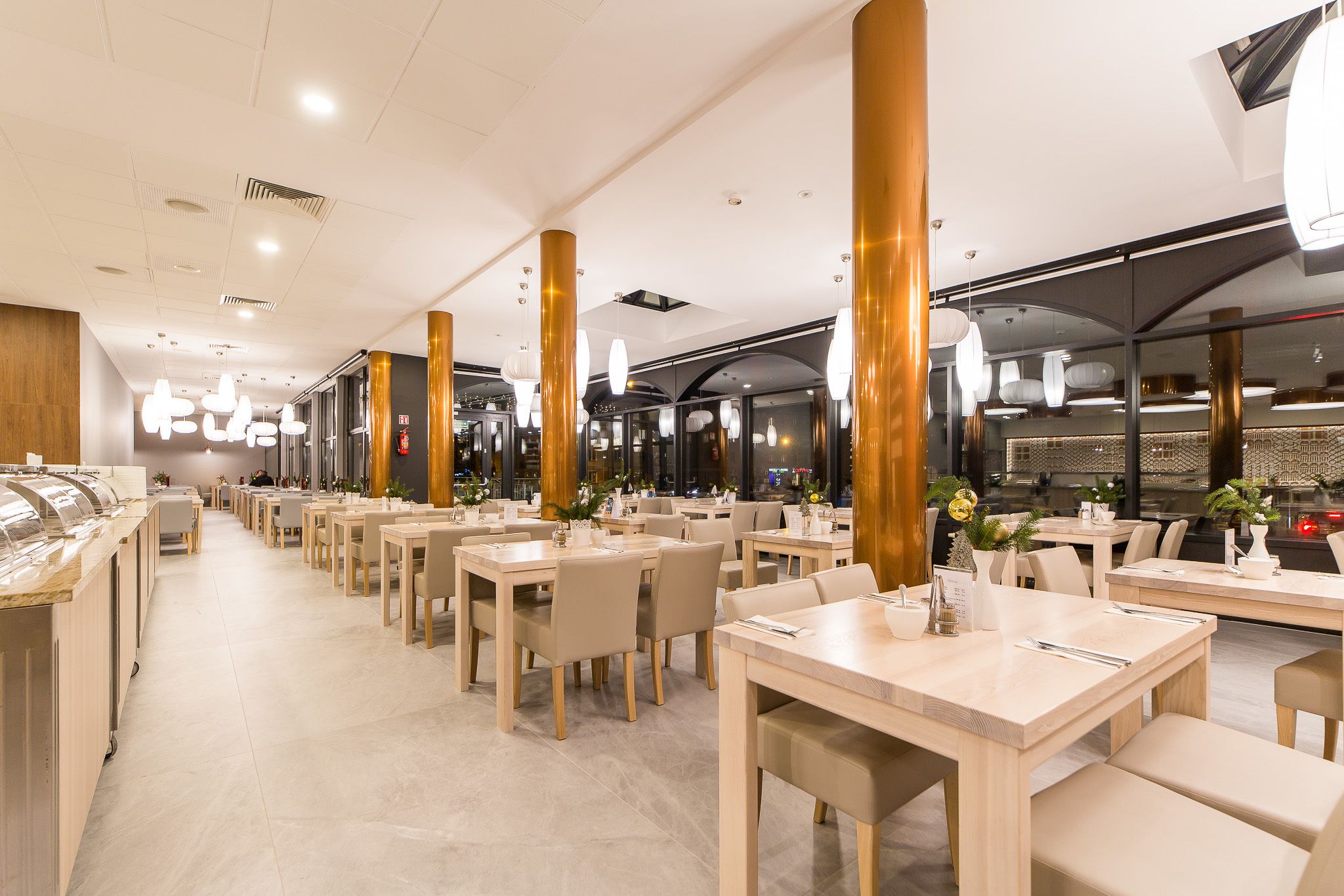 Un restaurant spacieux, doté de nombreuses tables et chaises, de grandes colonnes dorées et d’un éclairage moderne suspendu au plafond.