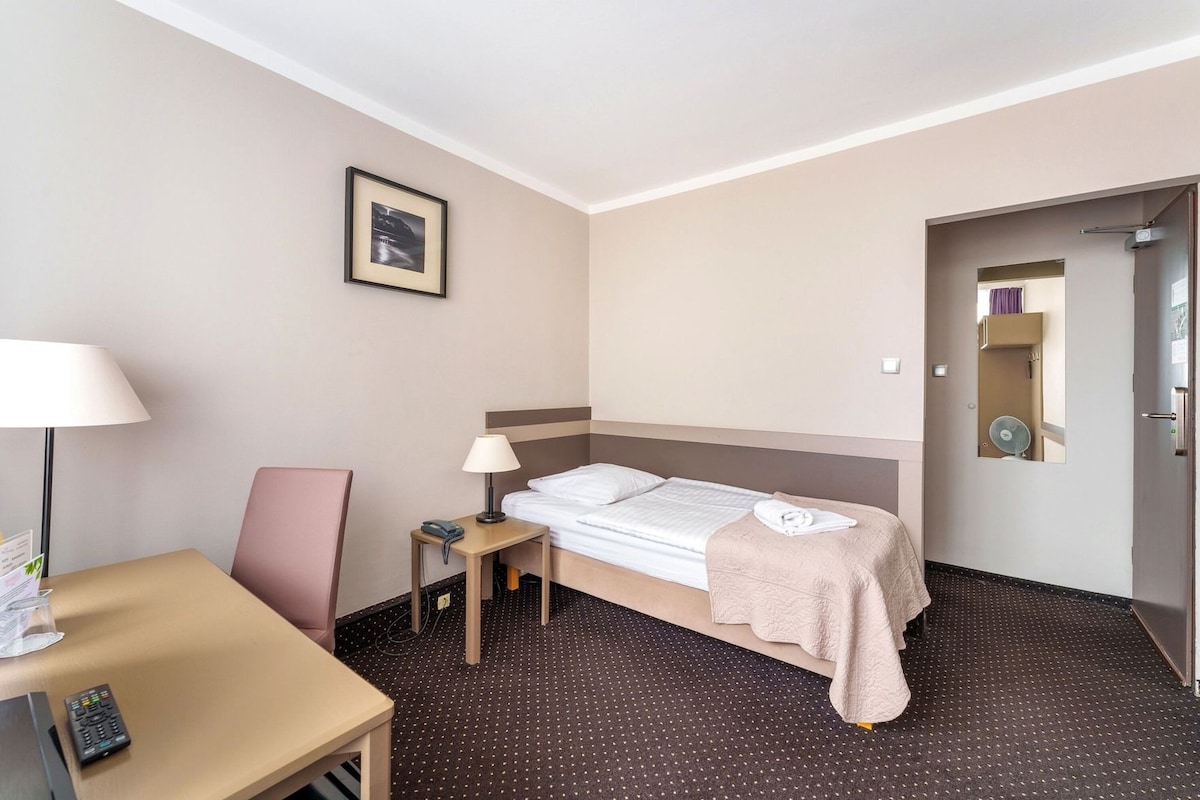 A hotel room with a bed, a desk, a chair, a lamp, a telephone, and a picture on the wall.