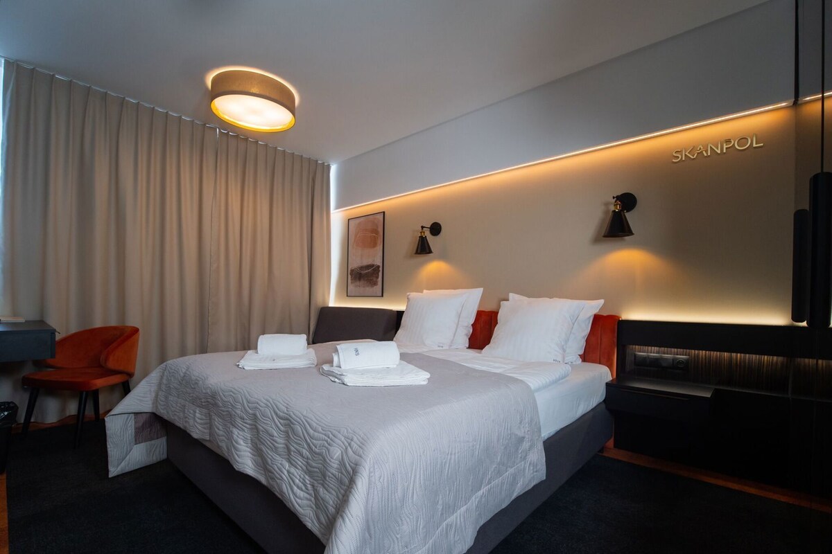 A hotel room with a bed, a desk, an orange chair, and a wall-mounted TV.
