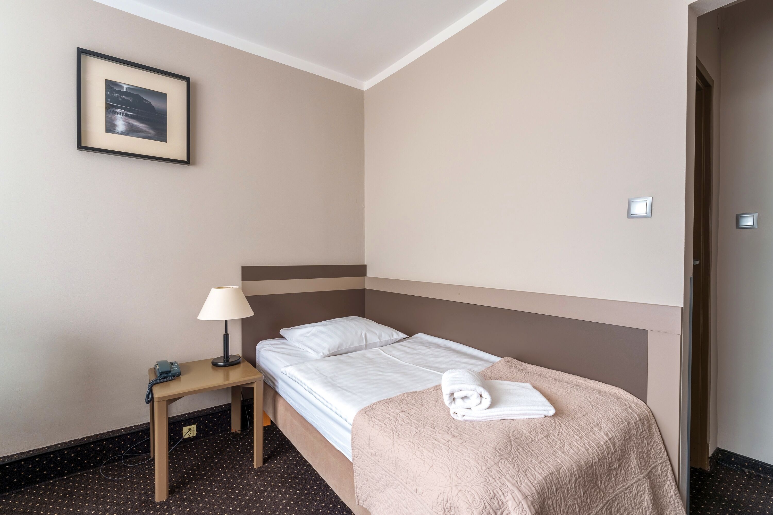 A hotel room with a single bed, a bedside table with a lamp, a framed picture on the wall, and a door to the right.