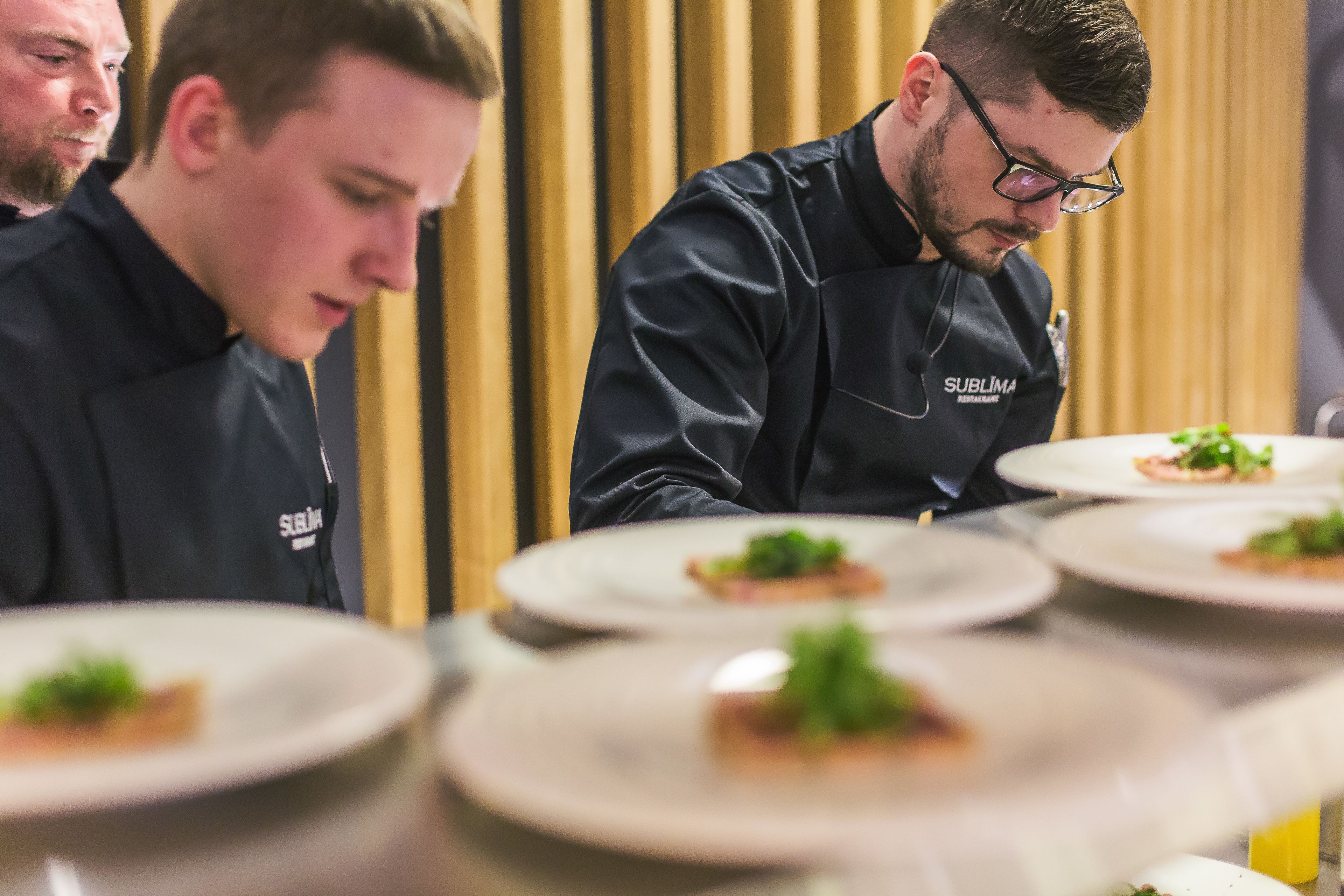 Köche in schwarzen Uniformen mit dem Schriftzug 'Sublima Restaurant' auf ihren Jacken bereiten Gerichte auf Tellern zu.