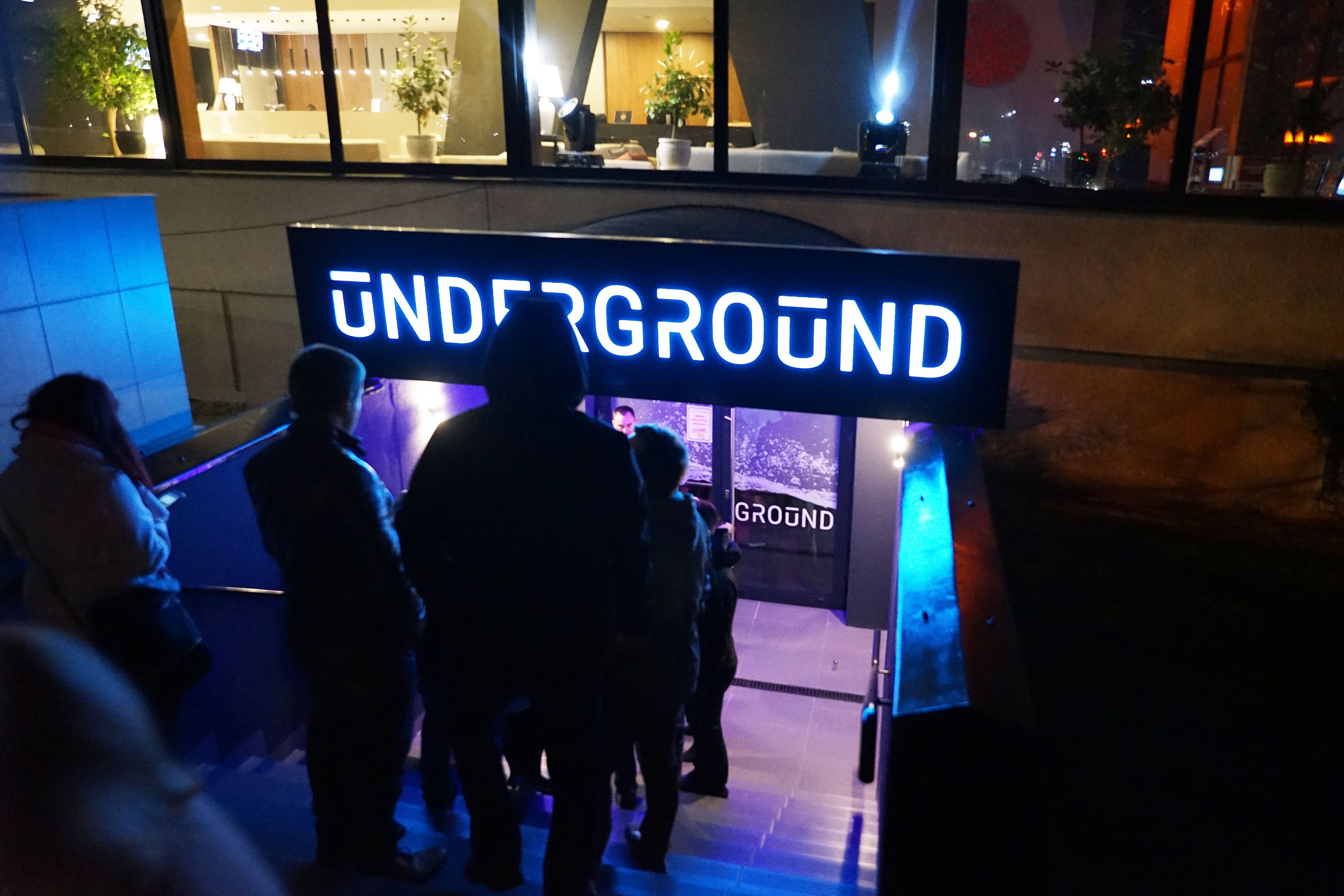 En skylt med texten ”UNDERGROUND” i en modern design, placerad i en stadsmiljö med tydliga arkitektoniska detaljer och en speciell atmosfär under kvälls- och nattetid.
