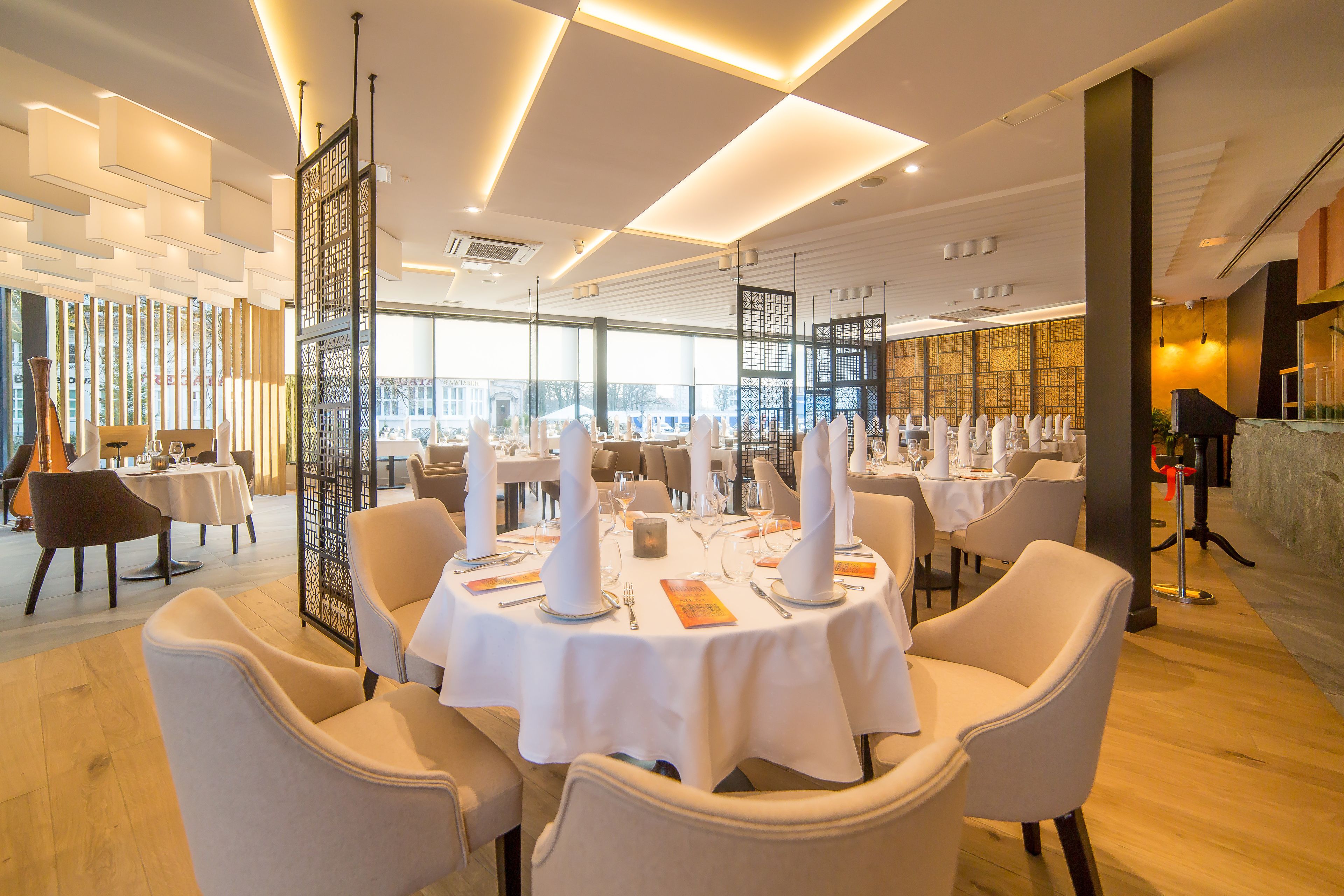 A modern restaurant with round tables set for dining, featuring a high ceiling with recessed lighting and a view of the cityscape through large windows.