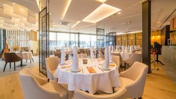 A modern restaurant with round tables set for dining, featuring a high ceiling with recessed lighting and a view of the cityscape through large windows.