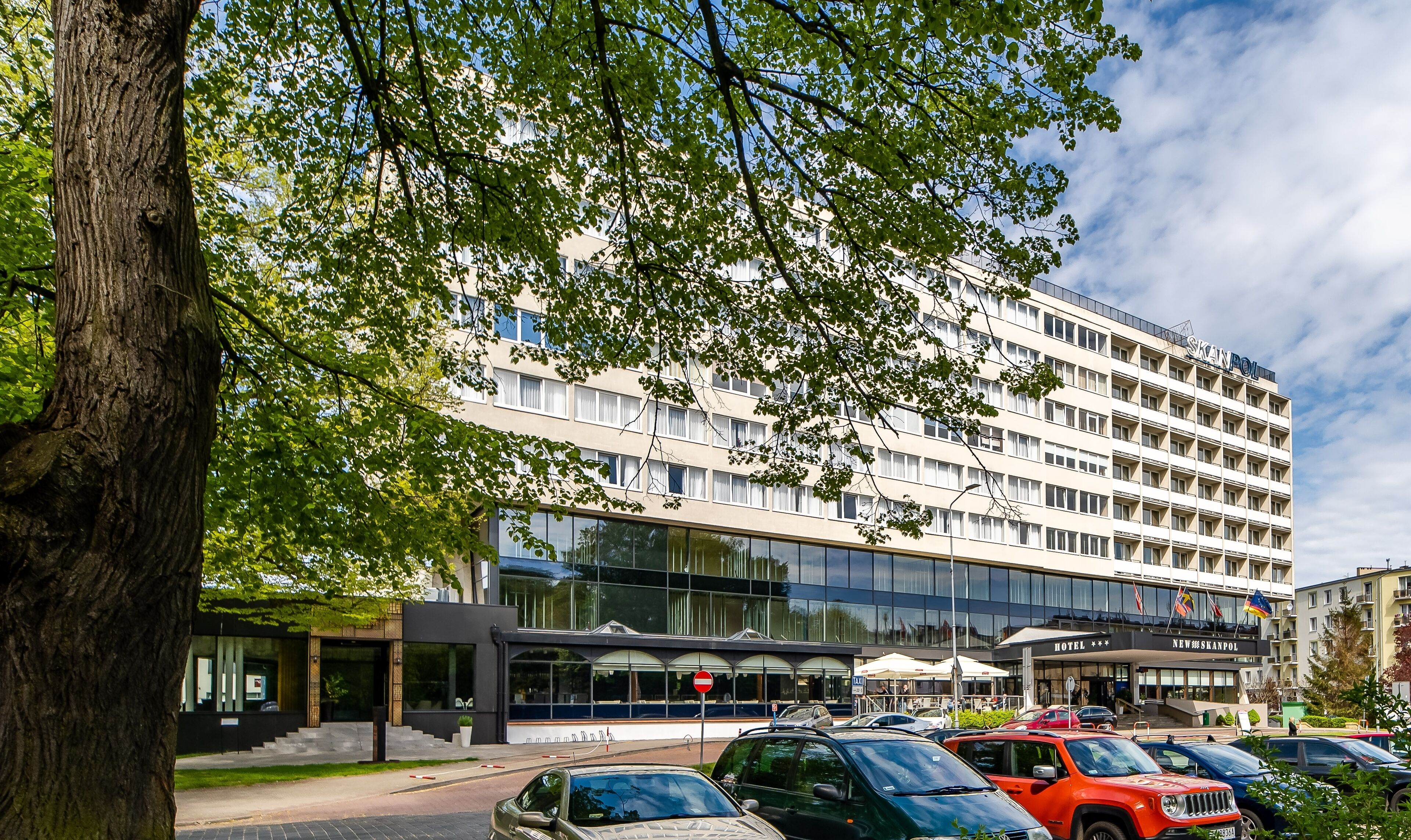 Un hotel de varios pisos con fachada moderna, amplias ventanas y una entrada visible con un cartel que dice 'HOTEL NEW SKANPOL'.