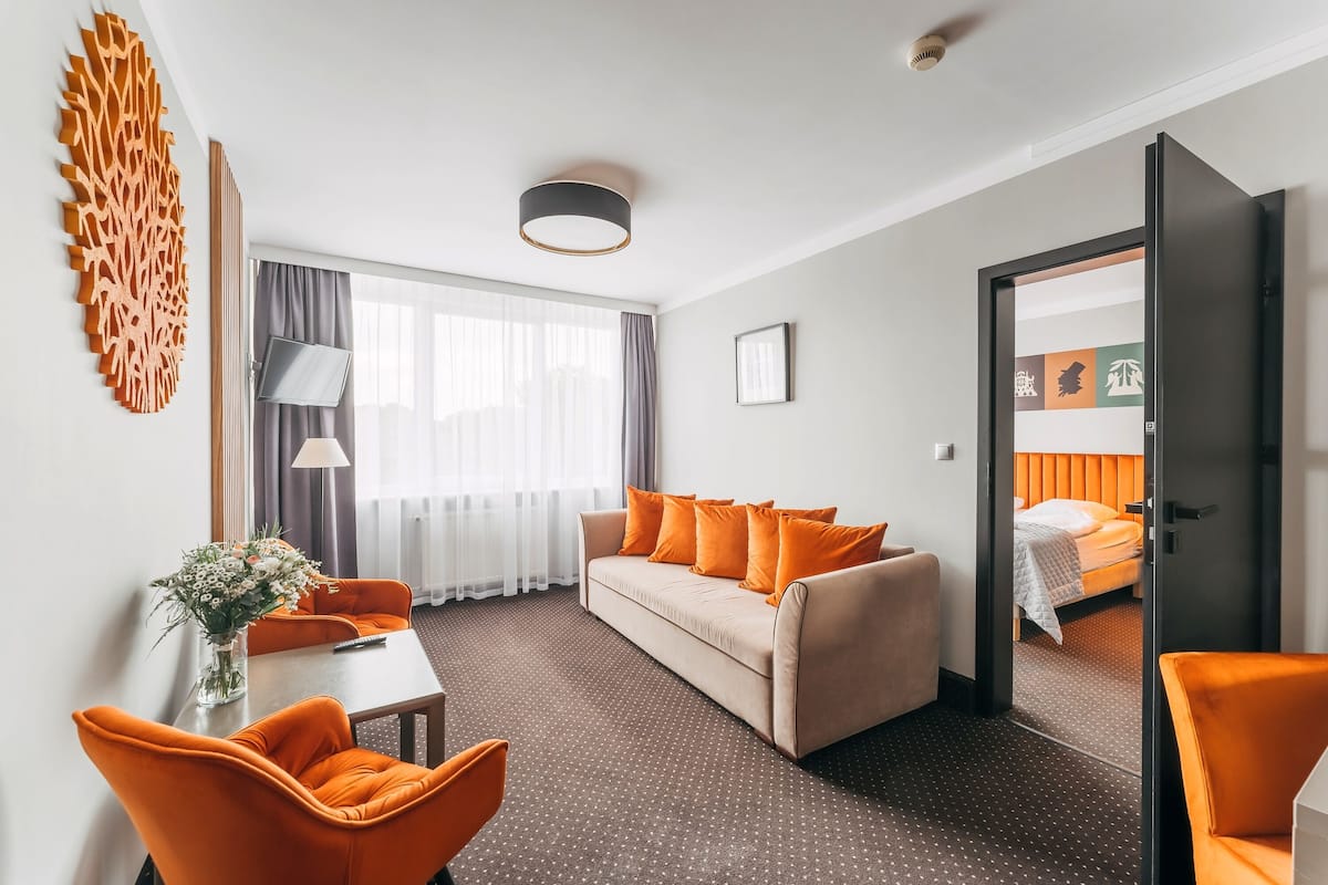 Une chambre d’hôtel avec un canapé, des fauteuils orange, une table à manger agrémentée d’un vase de fleurs, une télévision et un lit avec une tête de lit orange.