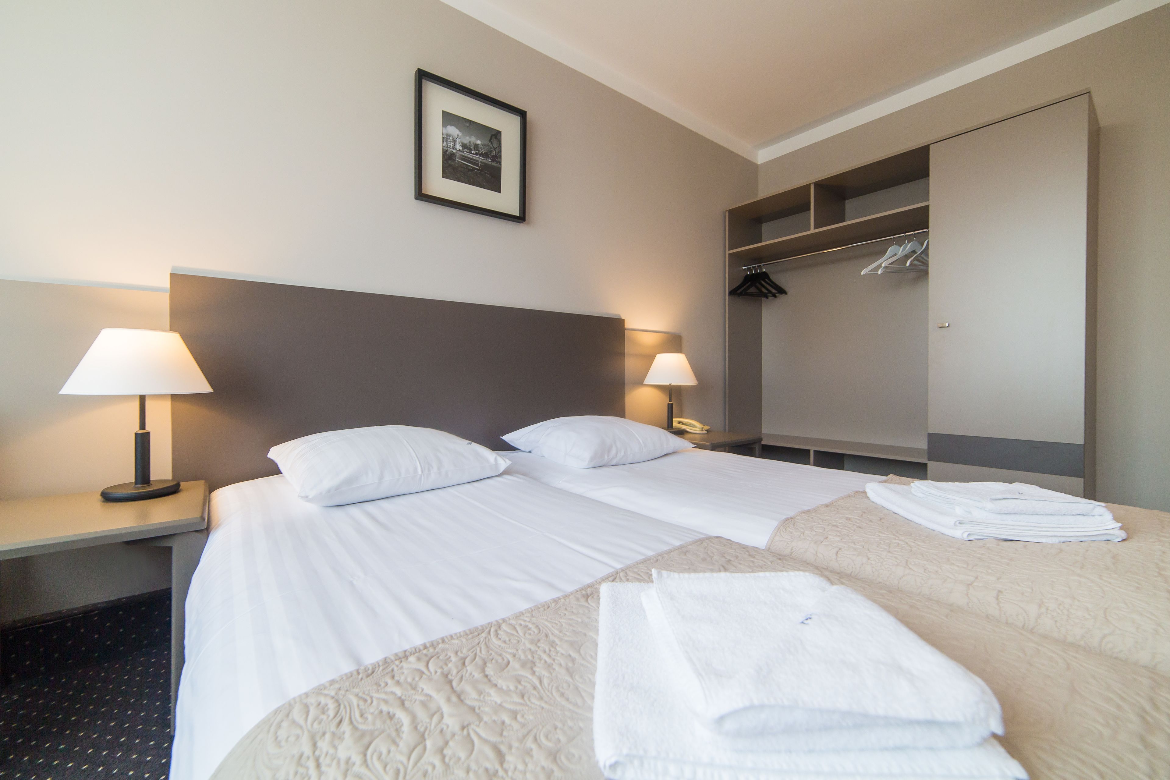 Habitación de hotel con dos camas, una mesita de noche con lámparas, un armario y un cuadro en la pared.
