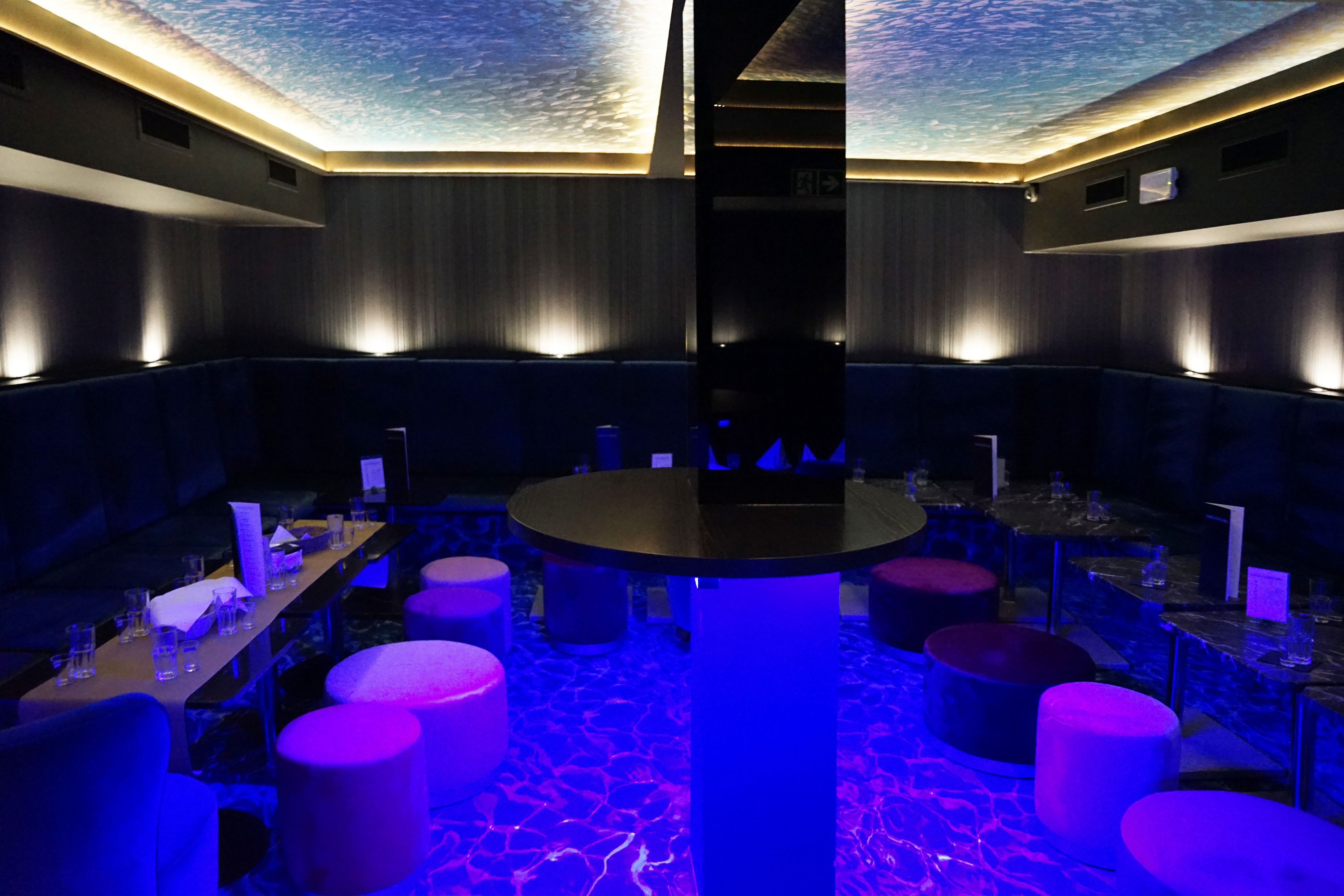 A modern lounge area with a central round table, multiple seating options, and a ceiling with a unique light pattern.