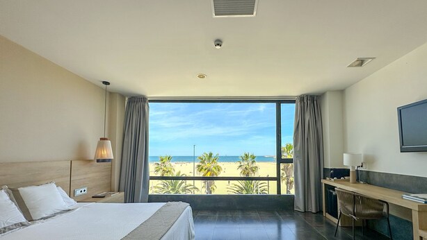 A hotel room with a bed, desk, and large window overlooking a beach. - El Coso Hotel (Valencia)
