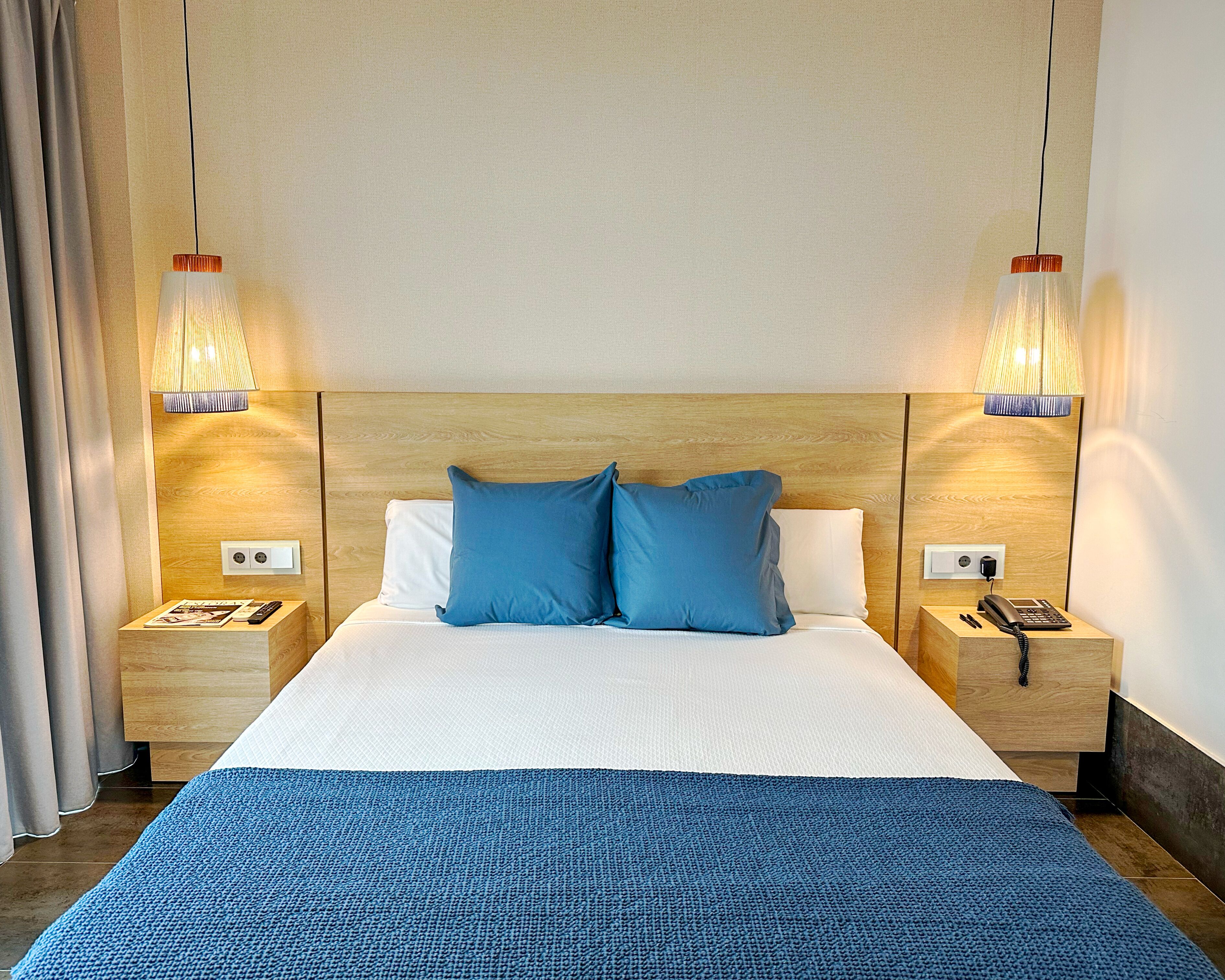 A hotel room with a large bed, two bedside tables with lamps, a wooden headboard, and a blue throw blanket.