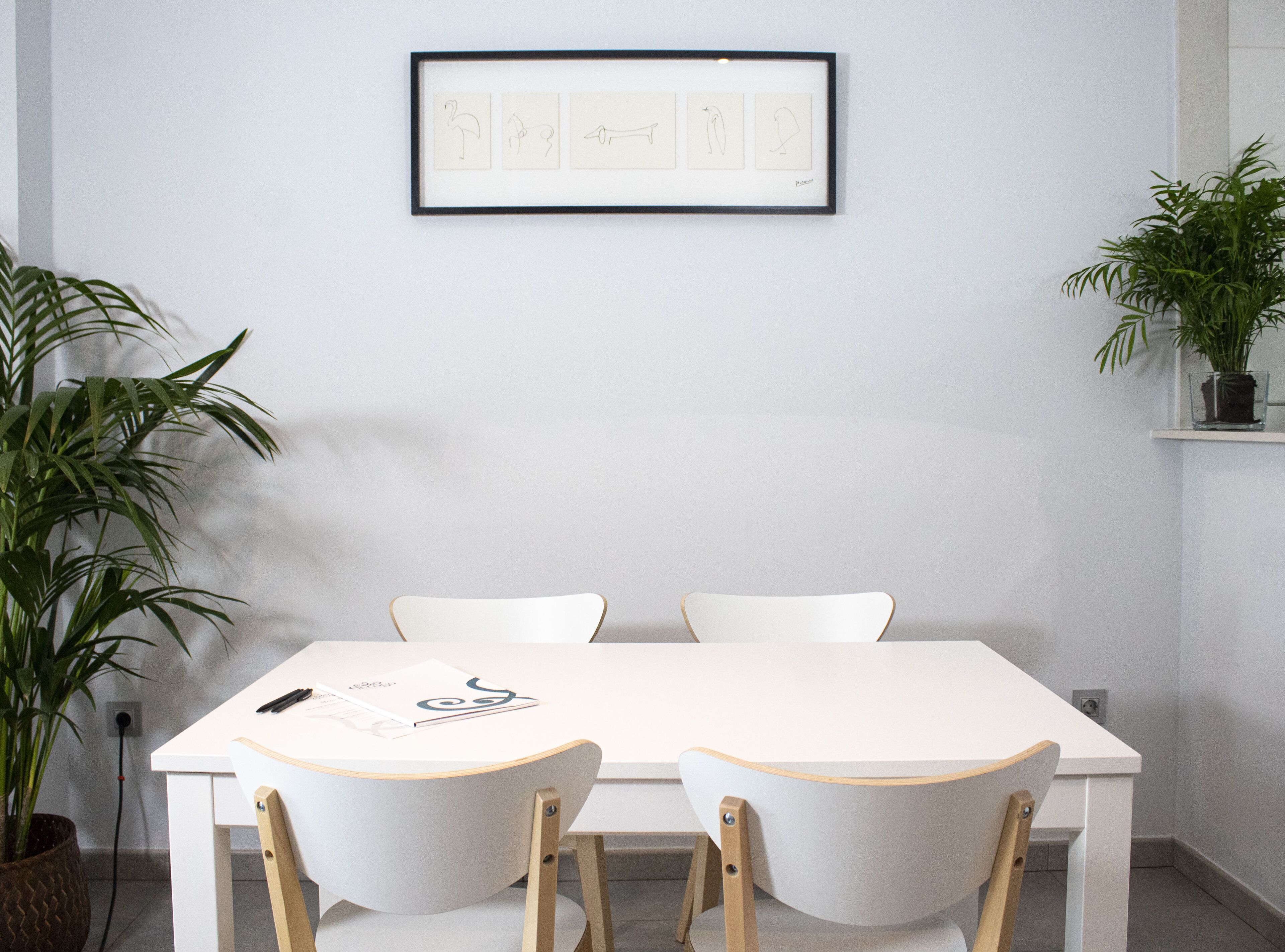 A minimalist dining area with a white table and chairs, framed artwork above, and potted plants on either side.