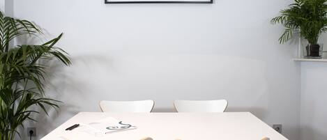 A minimalist dining area with a white table and chairs, framed artwork above, and potted plants on either side.