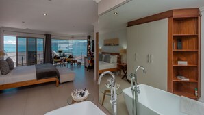 Un dormitorio moderno con una cama grande, un balcón con paredes de vidrio, una zona de estar con vistas y un baño separado con bañera y tocador.