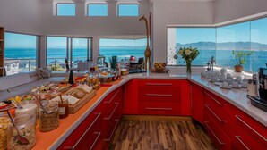 Una cocina moderna con una decoración roja, ventanas amplias con vistas al océano y una variedad de alimentos expuestos en la encimera.
