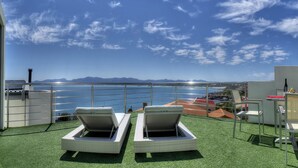 Un balcón con dos sillones de descanso, una mesa y una silla, con vista a un cuerpo de agua y montañas distantes.