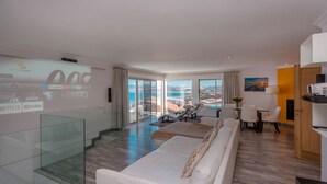 Una habitación de hotel moderna con balcón, sofá, zona de comedor y una pantalla de televisión que muestra servicios de streaming.