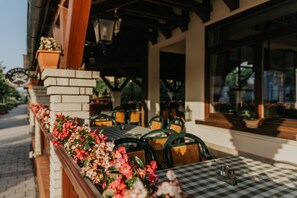 An outdoor seating area with tables and chairs, a brick planter with flowers, and a view of the street. - Korona Pension Restaurant (Hévíz)