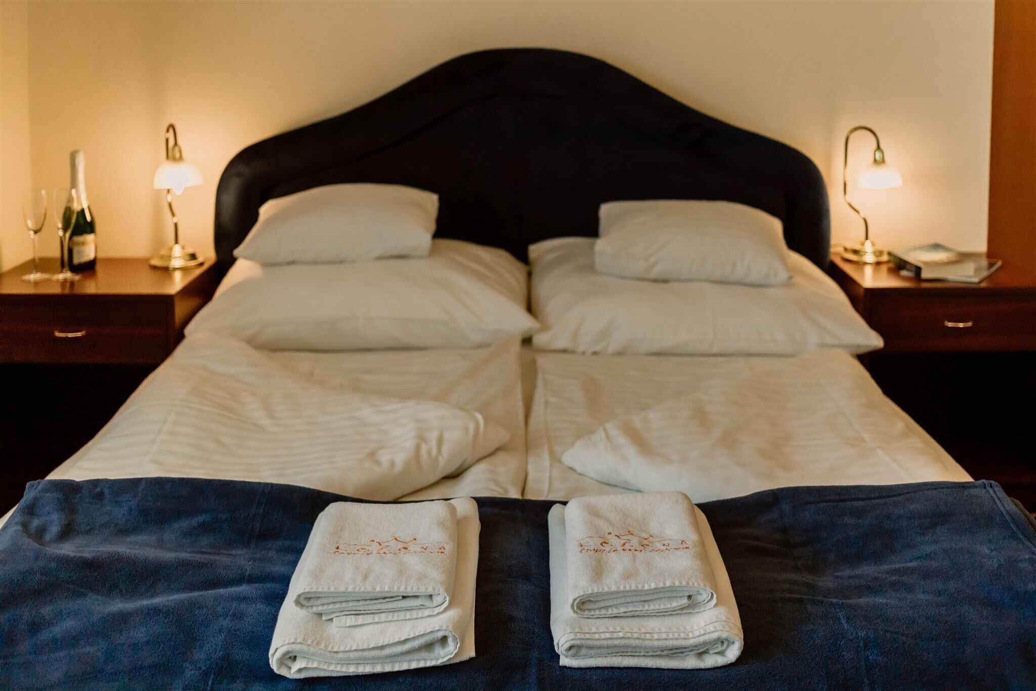 A double bed with white linens, two pillows, and two folded towels on a dark blue bedspread. There are two bedside tables with lamps and a bottle of champagne.